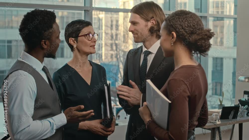 Medium shot of four professional multiethnic lawyers standing in contemporary office with big windows having conversation Medium shot of four professional multiethnic lawyers standing in contemporary office with big windows having conversation