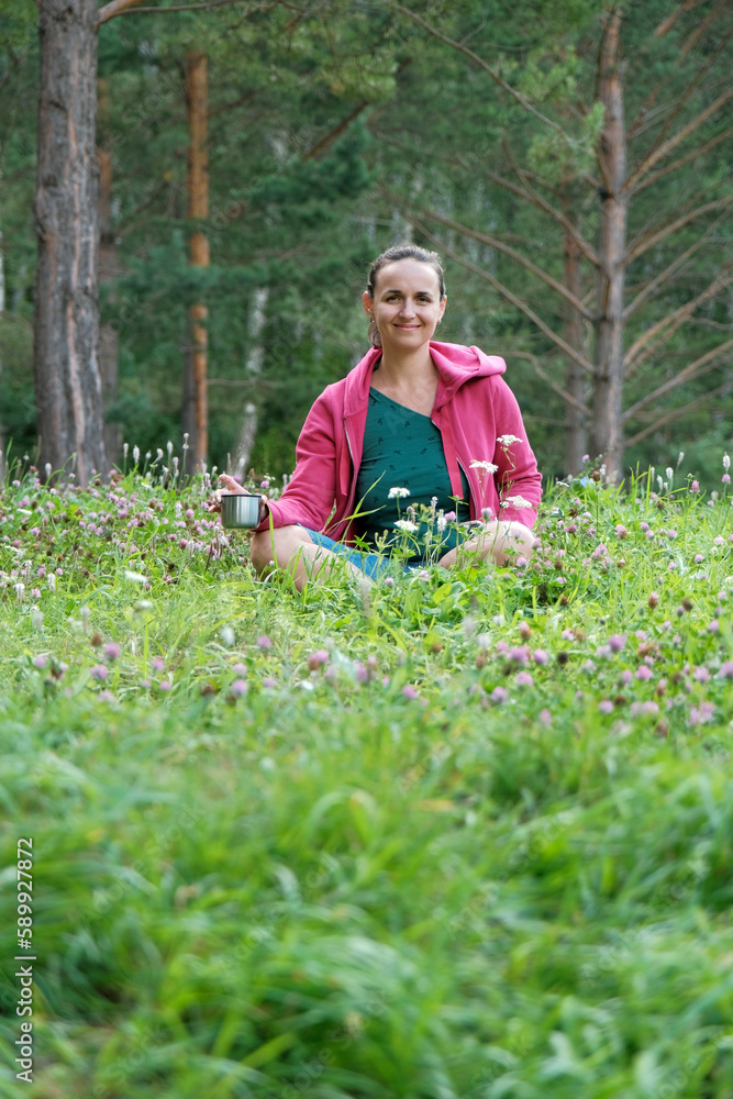 Smiling Young Woman Sitting on the Glade by the Pine Forest, Holding a Thermos Mug in Hands. Ecology and Harmony with Nature Concept 