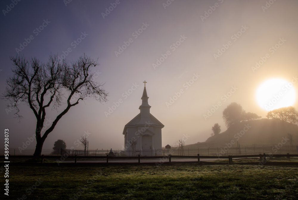 Bright sun and dark fog over small church and tree in rural countryside at dawn