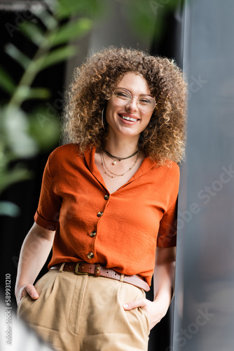 cheerful manager in glasses and hoop earrings standing with hands in pockets near wall in modern office.