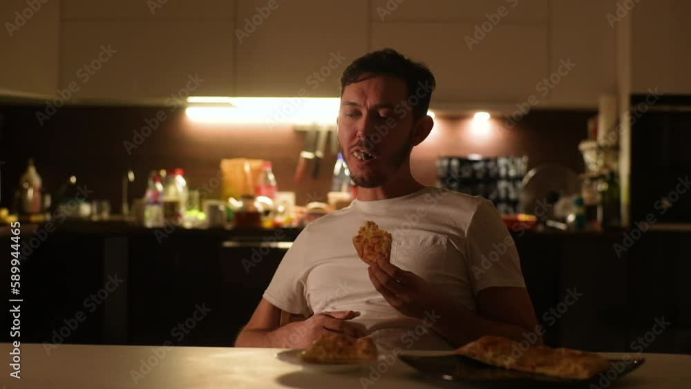 Portrait of depressed young man eating pizza sitting alone at table in evening or night on background of dark kitchen room. Front view of sad tired male eating junk food at home, slow motion. Portrait of depressed young man eating pizza sitting alone at table in evening or night on background of dark kitchen room. Front view of sad tired male eating junk food at home, slow motion.
