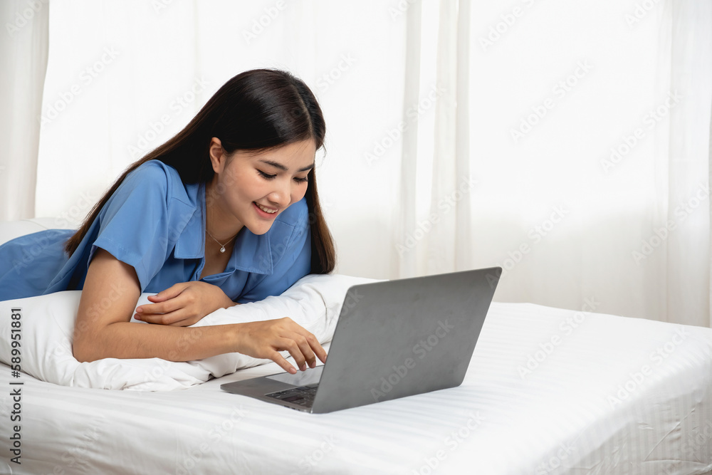 Young Asian woman with beautiful face, long hair, blue shirt lying on bed in white bedroom at home with cup of coffee and video call with laptop talking to relatives on vacation. holiday concept. Young Asian woman with beautiful face, long hair, blue shirt lying on bed in white bedroom at home with cup of coffee and video call with laptop talking to relatives on vacation. holiday concept.