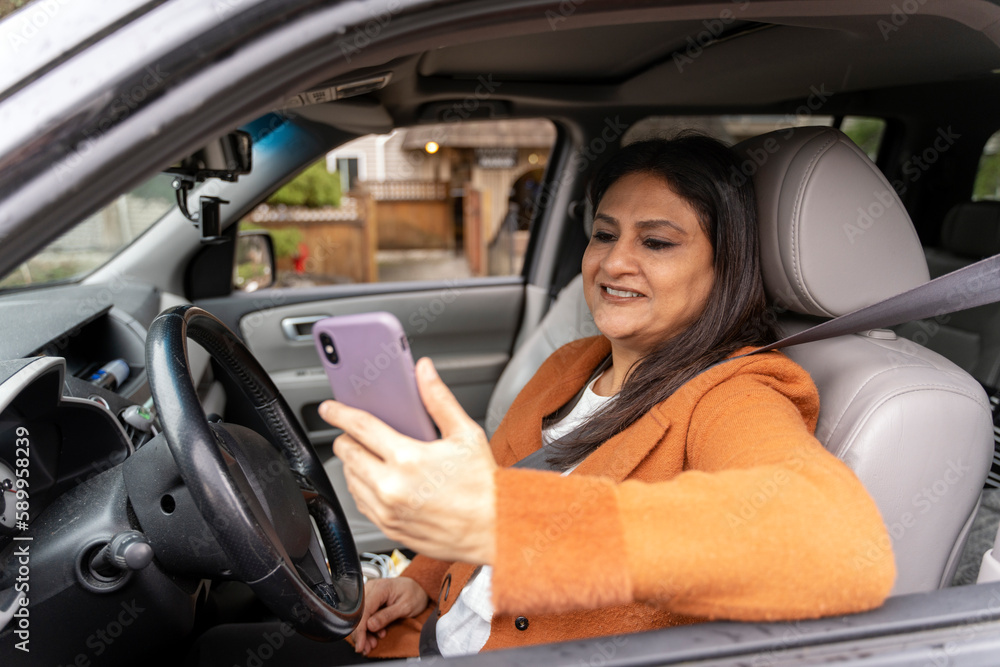 Confident smiling mature Indian woman holding mobile phone, checking email sitting inside new car. Transportation, distracted driving concept