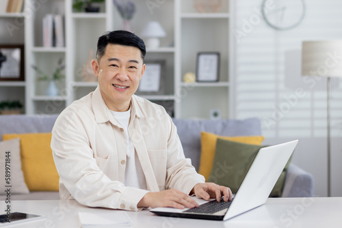 Portrait of successful Asian programmer, man working remotely from home office, businessman smiling and looking at camera using laptop at work sitting in living room at home.