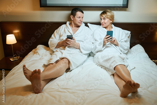 Man and woman in bathrobes are sitting on large bed