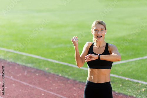 Sporty young woman athlete standing in the stadium wearing headphones and smiling at the camera, checking the time on the fitness bracelet, showing a victory gesture with her hand. © Liubomir