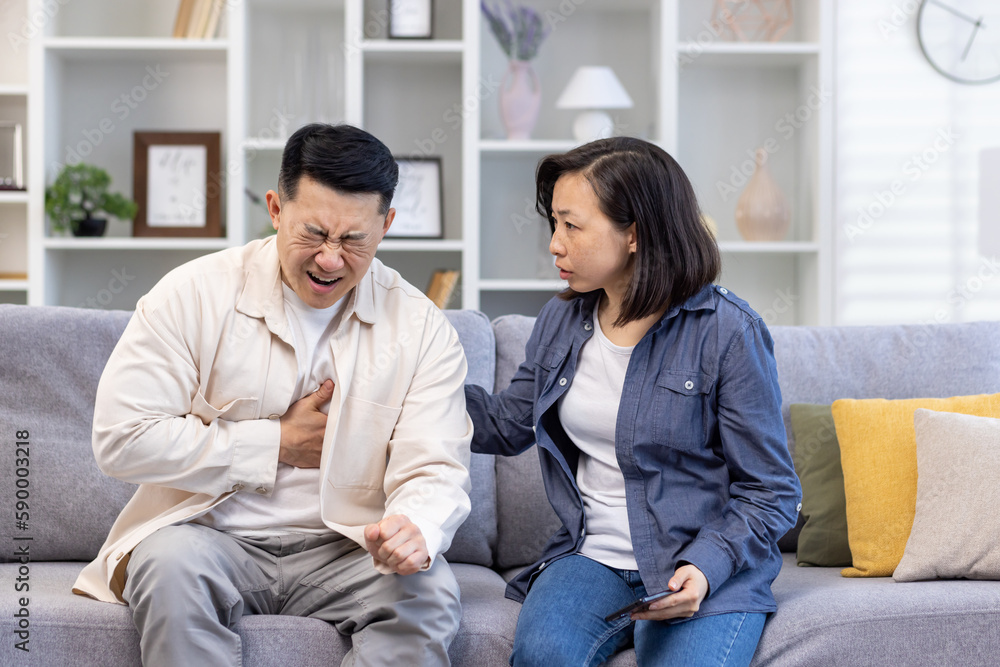Young Asian family sitting on sofa at home. A man holds his hand to his chest, heart attack, stroke, panic attack, feels severe pain. Shocked woman tries to help, give first aid. Young Asian family sitting on sofa at home. A man holds his hand to his chest, heart attack, stroke, panic attack, feels severe pain. Shocked woman tries to help, give first aid.