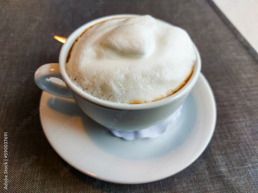 View of a mug of a cappuccino coffee with fluffy, white milk froth on the table covered with neutral grey tablecloth in front of window. Hot drink in a Café View of a mug of a cappuccino coffee with fluffy, white milk froth on the table covered with neutral grey tablecloth in front of window. Hot drink in a Café