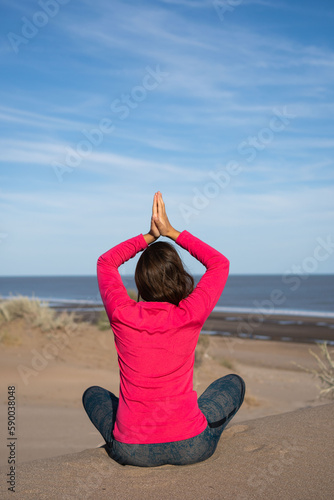 A woman sitting doing yoga mountain pose on the dunes overlooking the sea in Buenos Aires province, Argentina. Dressed in a pink thermal T-shirt. Vertical photo © Lautaro
