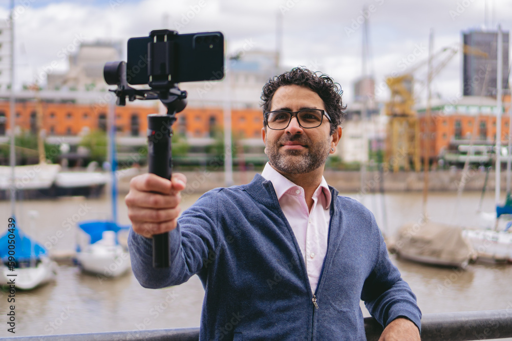 Middle-aged Latin man with glasses documenting with his cell phone a river port on a levee in the middle of the city. Middle-aged Latin man with glasses documenting with his cell phone a river port on a levee in the middle of the city.