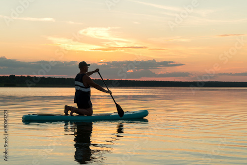 A man in shorts on his knees on a sapboard with an oar at sunset swims in the water of the lake against the backdrop of a pink sky reflection in the water.