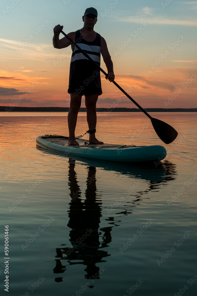 A man in shorts stands on a SUP board in the evening at sunset in the lake.