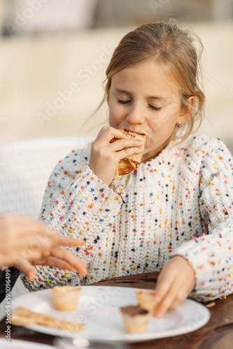 Cute little girl eating tasty crepes in cafe outdoors feeling satisfied and hungry with closed eyes