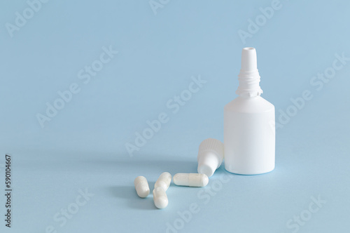 A bottle of nasal spray with the cap removed and some medication capsules placed next to it. White products on blue background. Horizontal.