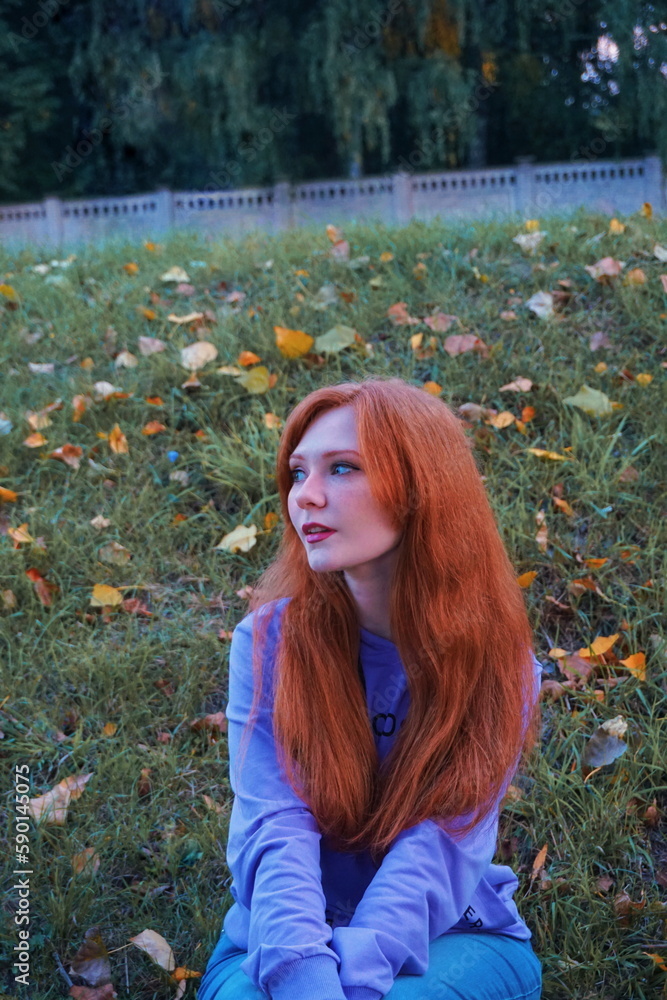 Naklejka premium red haired girl sits on the grass near the fallen leaves