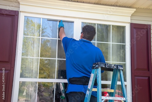 A Caucasian man on a ladder wearing blue latex gloves and listening to ear buds washing a window with a squeegee