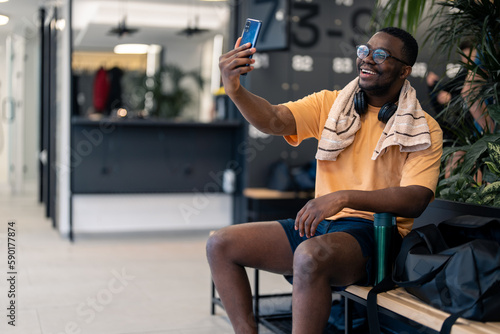 Handsome confident fit sports man sitting on bench in modern locker room with headphones and towel around his neck using mobile phone for video call at gym after successful workout training session.