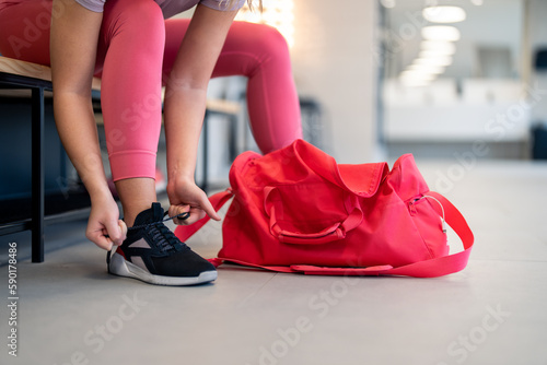 Cropped shot of fit sports woman in sportswear with gym bag wearing pink yoga pants and sneakers sitting on bench, getting ready for exercise session, tying her shoelaces in locker room at gym.