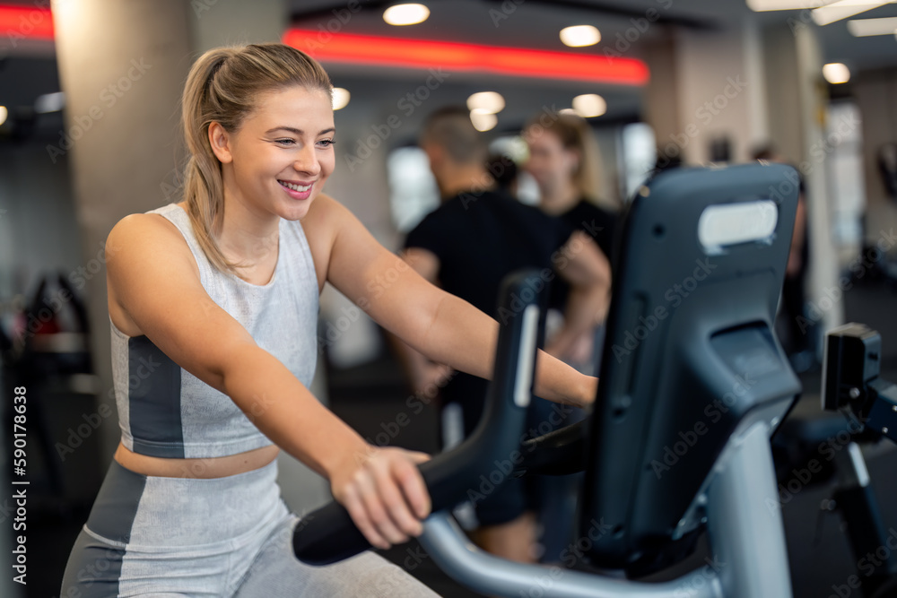 Young beautiful female gym member exercising at gym, having cardio training, cycling on stationary exercise bike, looking at screen of gym machine, improving endurance, practicing healthy lifestyle. Young beautiful female gym member exercising at gym, having cardio training, cycling on stationary exercise bike, looking at screen of gym machine, improving endurance, practicing healthy lifestyle.