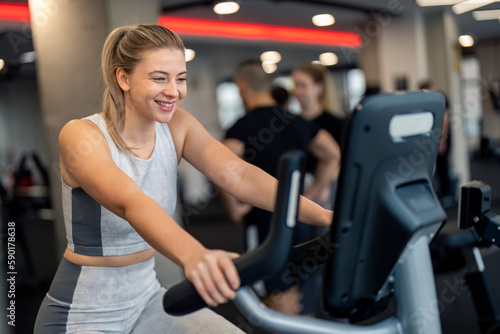 Young beautiful female gym member exercising at gym, having cardio training, cycling on stationary exercise bike, looking at screen of gym machine, improving endurance, practicing healthy lifestyle.