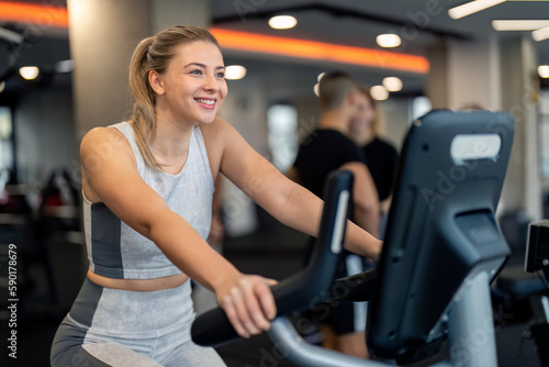Young sportswoman athlete wearing sportswear exercising at gym, feeling positive and motivated, having cardio training day, cycling on exercise bike, improving endurance, keeping her body in shape.