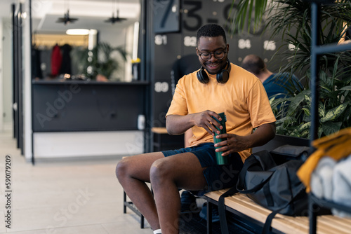 Young athlete man sportsperson in yellow t-shirt with eyeglasses and headphones holding opening water bottle sitting on bench in locker room at gym with smile after completed workout training session.