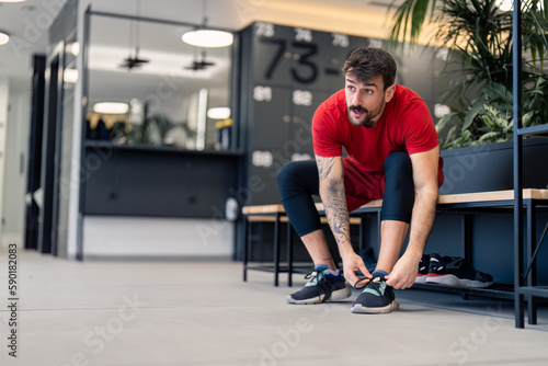 Handsome sportsman fitness coach or gym client with tattoos in a hurry sitting on bench tying fast his shoelaces in modern locker room worried about getting late on exercise workout session in gym.
