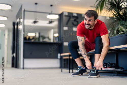 Handsome confident fit sportsman gym coach with tattoos sitting on bench tying his shoelaces in modern locker room getting ready for workout training session to keep his body healthy and in shape.