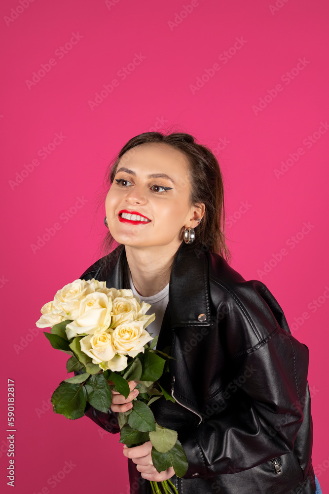 Cool biker girl wearing a leather jacket and jeans posing with a bouquet of white roses. Isolated on pink background. Cool biker girl wearing a leather jacket and jeans posing with a bouquet of white roses. Isolated on pink background.