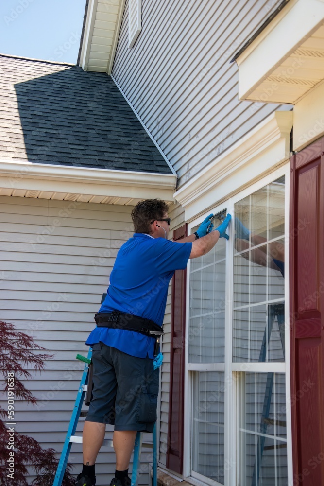 A Caucasian man on a ladder wearing blue latex gloves and listening to ear buds washing a window with a squeegee A Caucasian man on a ladder wearing blue latex gloves and listening to ear buds washing a window with a squeegee
