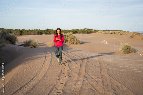 Young bespectacled Latina woman walking face forward using her cell phone on the dunes of a South American beach in Buenos Aires province. © Lautaro
