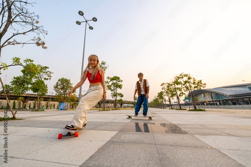 Fototapeta premium Group of Young Asian man and woman friends skating on longboard skate at public park on summer vacation. Stylish people have fun urban lifestyle practicing outdoor sport skateboarding at city street.
