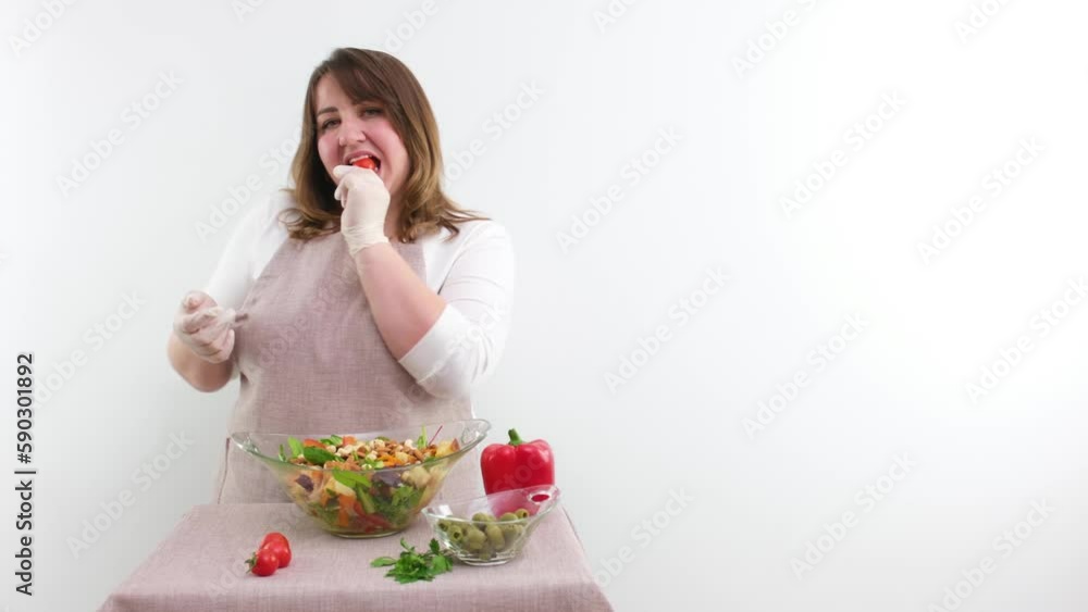 plump woman chef tosses cherry tomato sexy eats it smiles makes eyes on a white background space for advertising text cleanliness tidiness aprons gloves shows thumb up class preparing a salad