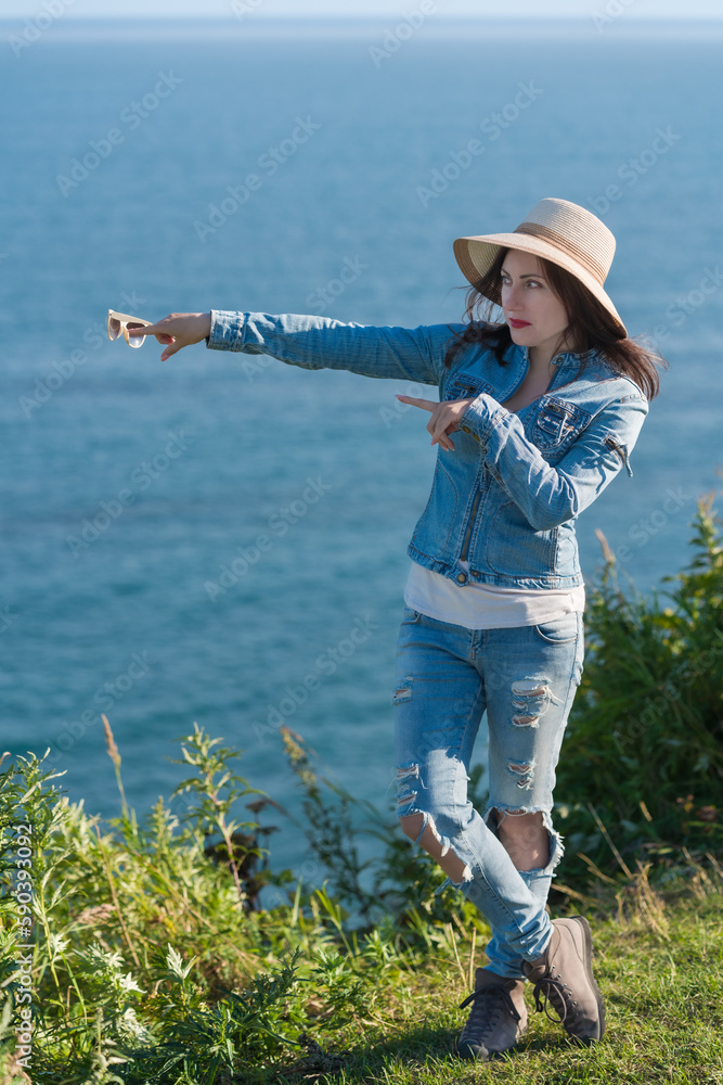 Woman in hat, denim jacket and jeans shows hands direction to side against backdrop of sky and sea