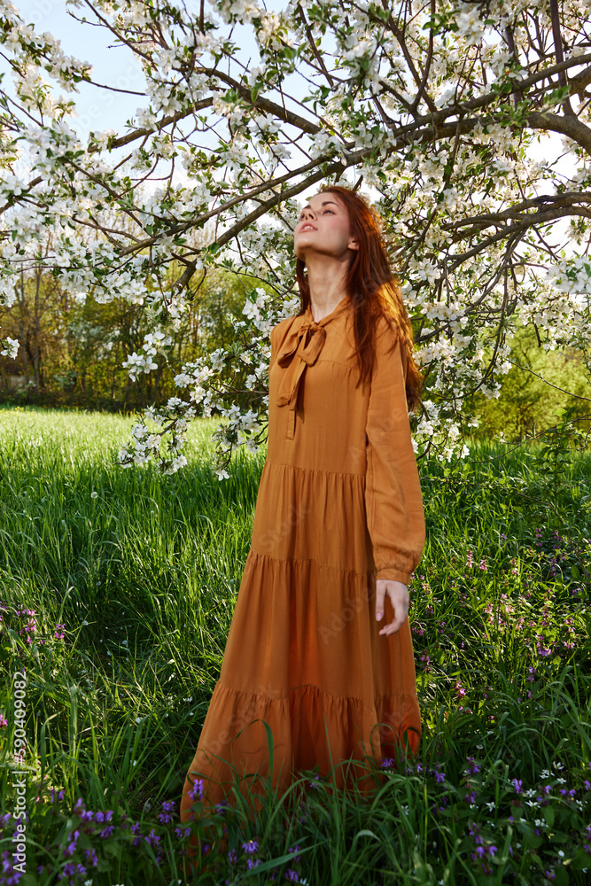 a happy, modest woman stands in an orange dress near a flowering tree and enjoys nature and a sunny summer day a happy, modest woman stands in an orange dress near a flowering tree and enjoys nature and a sunny summer day