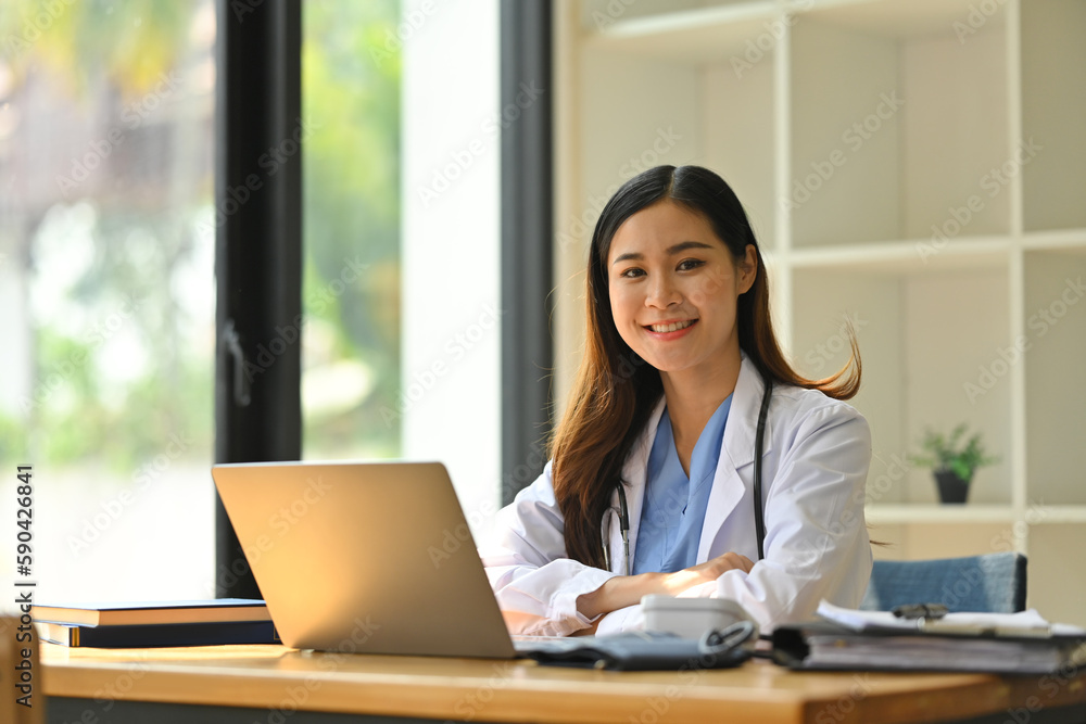 Confident asian female doctor in white coat and stethoscope using laptop computer, sitting in medical office Confident asian female doctor in white coat and stethoscope using laptop computer, sitting in medical office