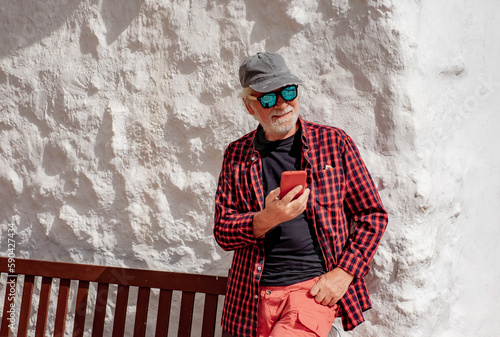 Smiling Senior Man in Checkered Red Shirt Standing Outdoors Against a While Wall Using Mobile Phone