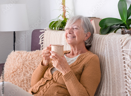 Smiling Senior Woman Sitting Indoors in Cozy Sofa Holding a Coffee Cup. Elderly Lady Having Relaxing Moments at Home