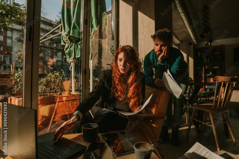 Gorgeous ginger businesswoman working on her lap top while holding paperwork in the other hand. She is explaining something to her male coworker Gorgeous ginger businesswoman working on her lap top while holding paperwork in the other hand. She is explaining something to her male coworker