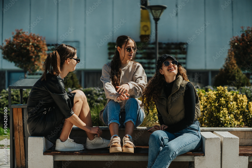 Fototapeta premium Three lovely stylish girls sitting on a bench and having a fun conversation, on a beautiful sunny day