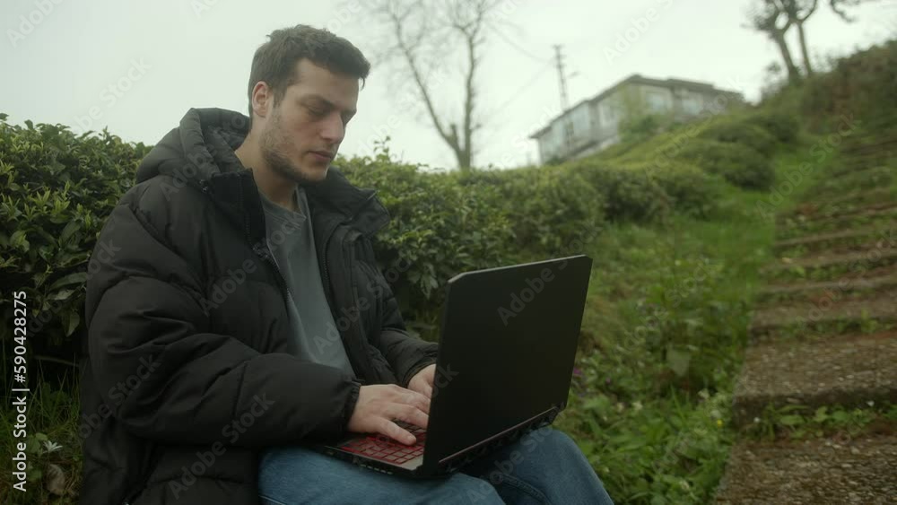 Handsome young man with brown hair and black jacket using his laptop on a beautiful nature background Handsome young man with brown hair and black jacket using his laptop on a beautiful nature background