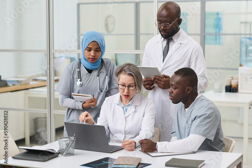 Doctor in white coat discussing difficult disease with her colleagues on laptop during teamwork in office