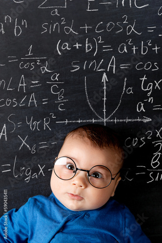 A beautiful little baby with glasses, a concept of learning and knowledge. Chalkboard with math formulas in the background.