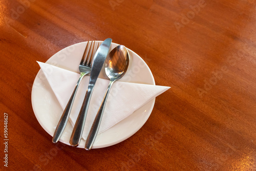 Spoon, fork and knife in white plate on wooden table waiting meal at restaurant.