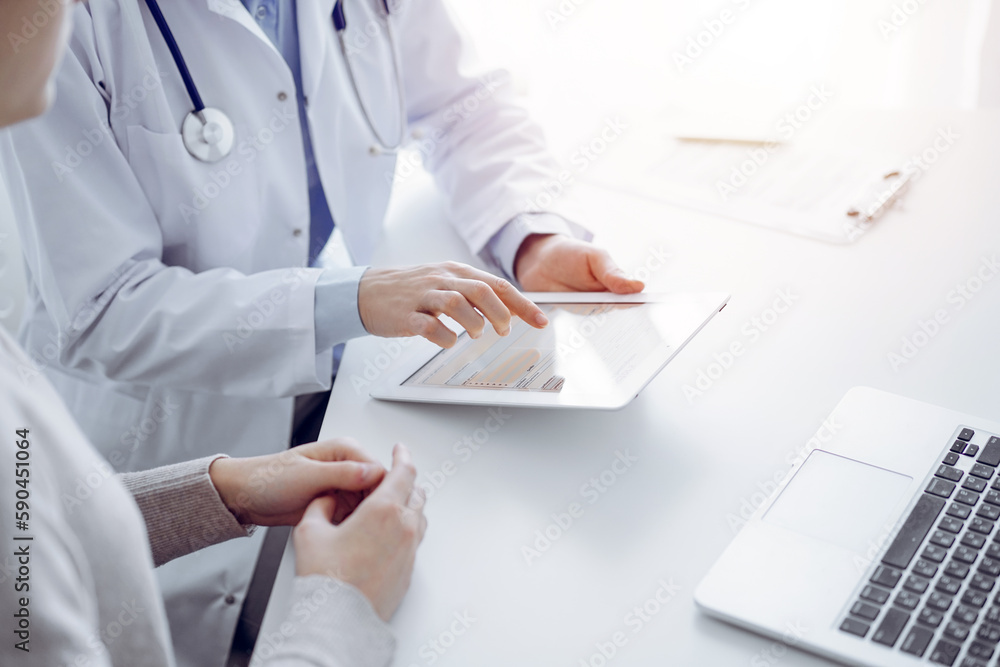 Doctor and patient sitting at the table in clinic. The focus is on female physician's hands using tablet computer, close up. Medicine and healthcare concept Doctor and patient sitting at the table in clinic. The focus is on female physician's hands using tablet computer, close up. Medicine and healthcare concept