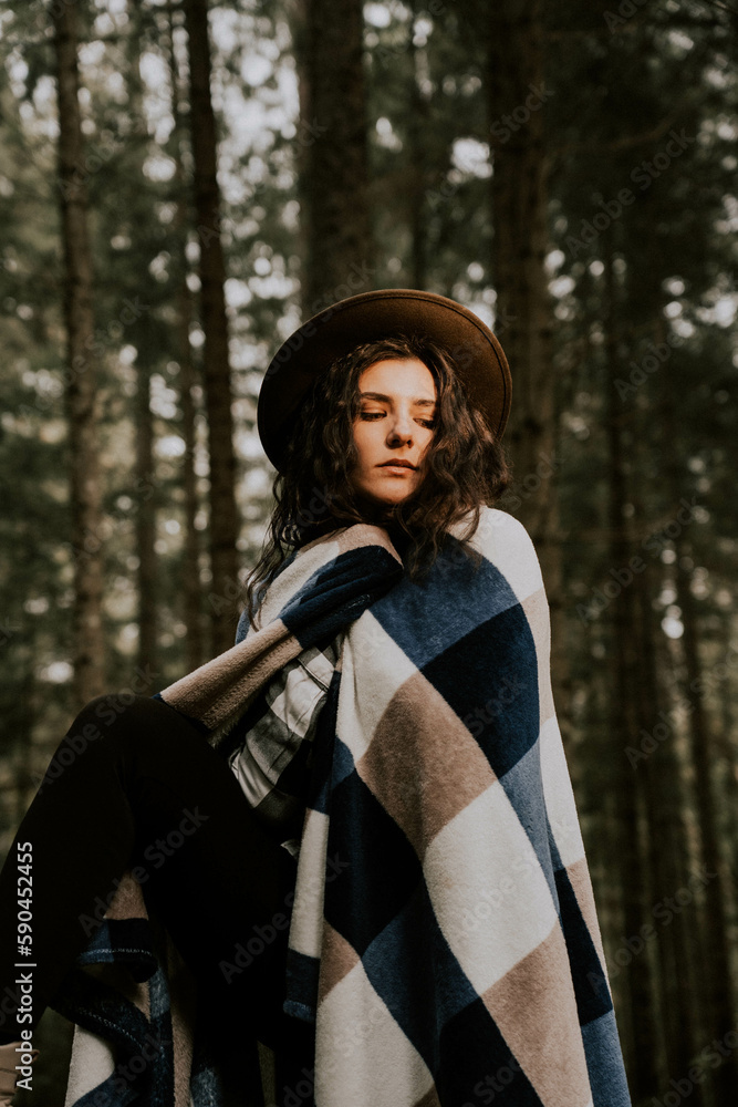 Stylish hipster girl in a hat wrapped in a checkered plaid posing in the base of a pine forest.Travel concept