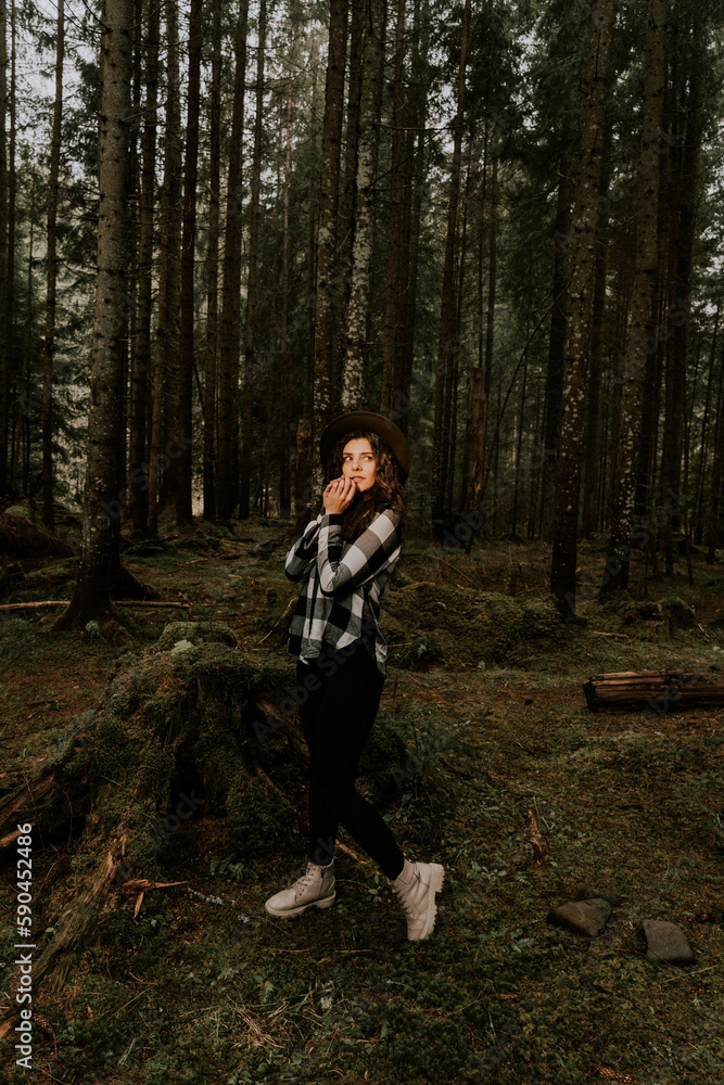 Stylish hipster girl in a hat wrapped in a checkered plaid posing in the base of a pine forest.Travel concept Stylish hipster girl in a hat wrapped in a checkered plaid posing in the base of a pine forest.Travel concept