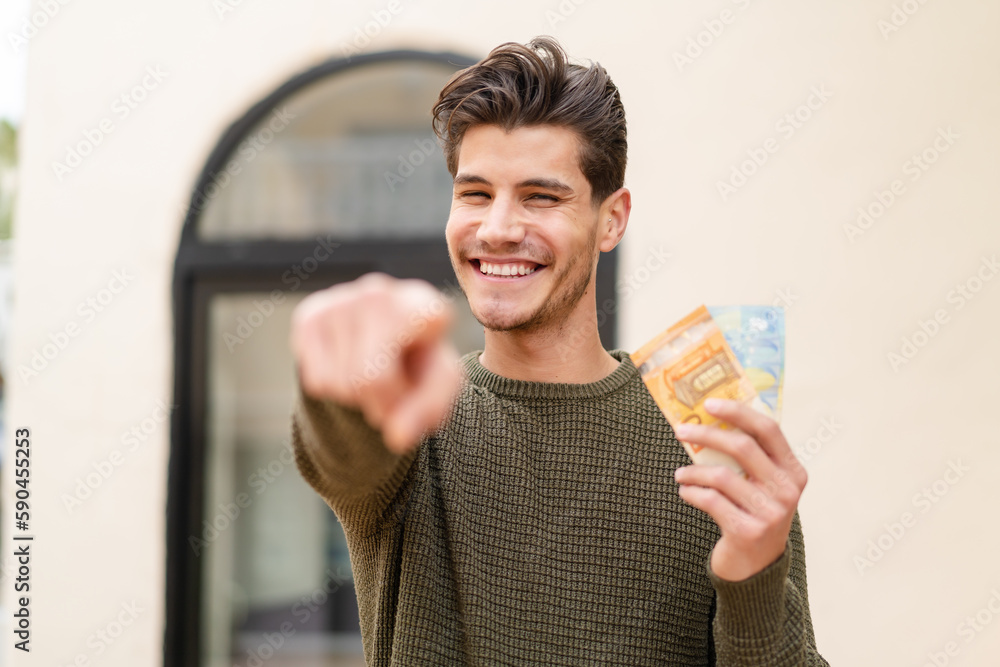 Young caucasian man taking a lot of money at outdoors points finger at you with a confident expression Young caucasian man taking a lot of money at outdoors points finger at you with a confident expression