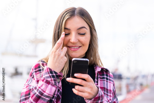 Young pretty Romanian woman at outdoors using mobile phone with fingers crossing