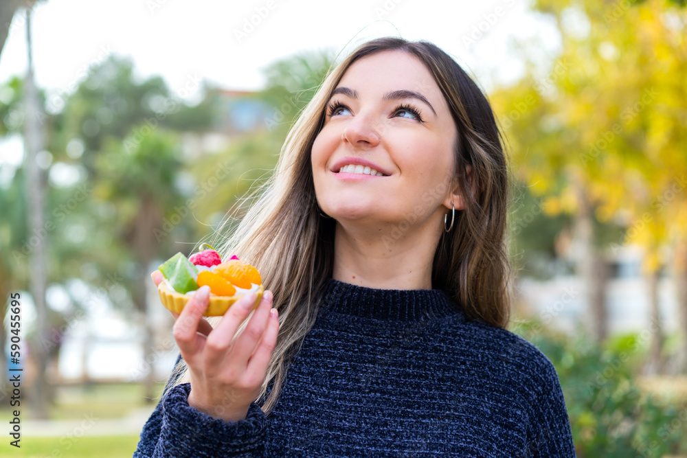 Young pretty Romanian woman holding a tartlet at outdoors looking up while smiling Young pretty Romanian woman holding a tartlet at outdoors looking up while smiling